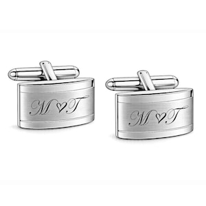 May include: A pair of silver-toned oval cufflinks with a brushed metal finish. Each cufflink features an engraved script monogram of the letters M and T separated by a small heart symbol. The cufflinks have a standard swivel toggle backing.