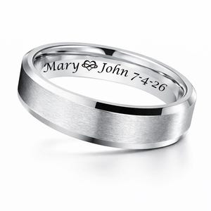 May include: A silver-colored ring with a brushed finish. The ring has the engraved names "Mary" and "John" with a heart symbol, and the date "7-4-26". The ring is likely a wedding band or a commemorative piece.