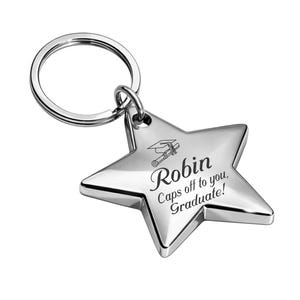 May include: A silver star-shaped keychain with a graduation cap and scroll design. The keychain has the name "Robin" and the text "Caps off to you, Graduate!" engraved on it. The keychain is attached to a silver ring.