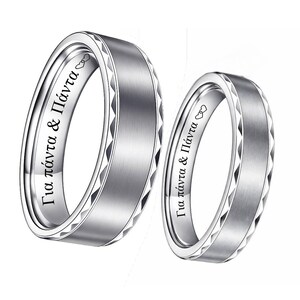May include: Two silver wedding bands. The bands have a brushed metal finish with a geometric pattern around the edges. The inside of each ring has Greek text and a heart symbol. The rings are displayed on a white background.