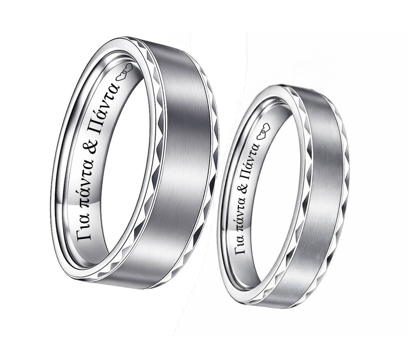 May include: Two silver wedding bands. The bands have a brushed metal finish with a geometric pattern around the edges. The inside of each ring has Greek text and a heart symbol. The rings are displayed on a white background.