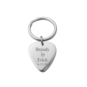 May include: A silver guitar pick-shaped keychain. The keychain has a split ring and a pendant engraved with the names "Brandy" and "Erick", a musical note, and the date "6-13-26". The keychain is made of a shiny, reflective metal.