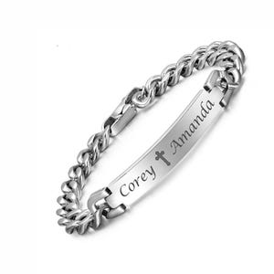 May include: A silver-toned metal curb chain bracelet featuring a central rectangular identification plate. The plate is engraved with the names Corey and Amanda, separated by a small cross symbol in a dark, legible script font.