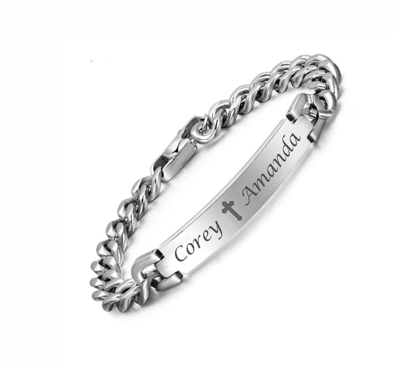 May include: A silver-toned metal curb chain bracelet featuring a central rectangular identification plate. The plate is engraved with the names Corey and Amanda, separated by a small cross symbol in a dark, legible script font.