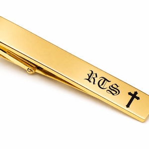 May include: A gold-colored tie bar with the initials "RTS" and a cross symbol engraved in black. The tie clip is rectangular and has a hinged clasp.