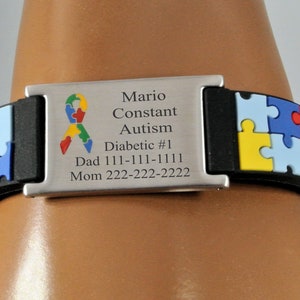 May include: A black and silver medical ID bracelet with a puzzle piece design. The bracelet has a silver plate with the words "Mario Constant, Autism, Diabetic #1" and contact information. The puzzle pieces are yellow, red, blue, and green.