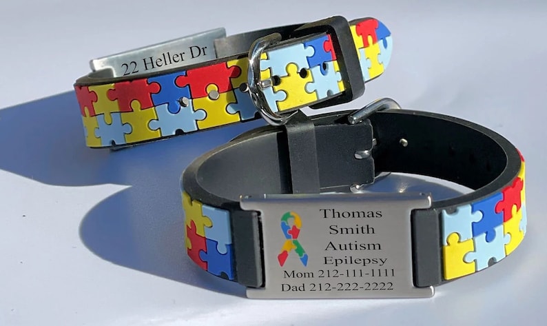 May include: Two black silicone bracelets with puzzle piece designs in red, yellow, blue, and green. One bracelet has the address "22 Heller Dr" engraved. The other has a medical alert tag with a ribbon symbol and the words "Autism" and "Epilepsy".