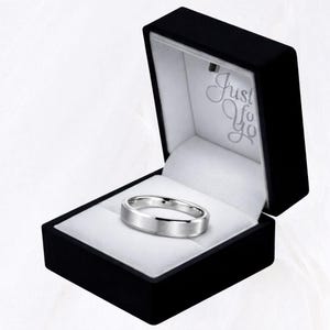 May include: A silver-colored ring with a brushed finish rests inside a black ring box. The box is open, revealing a white interior and the inscription "Just For You". The ring has a flat top and beveled edges.