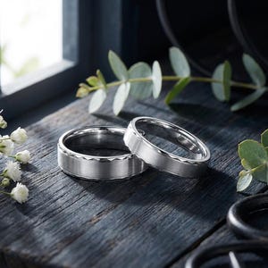 May include: Two silver wedding bands with a textured design on a dark wooden surface. The rings are displayed with greenery and white flowers, creating a romantic and elegant scene. The bands have a matte finish.