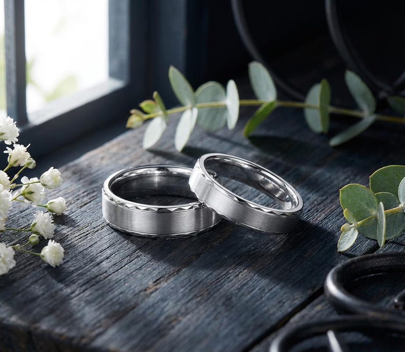 May include: Two silver wedding bands with a textured design on a dark wooden surface. The rings are displayed with greenery and white flowers, creating a romantic and elegant scene. The bands have a matte finish.