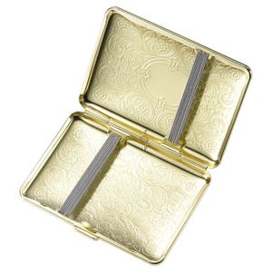 Puede incluir: Estuche de cigarrillos dorado con un dise&ntilde;o floral ornamentado. El estuche est&aacute; abierto y tiene dos bandas el&aacute;sticas para sujetar los cigarrillos.