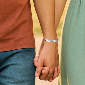 May include: A silver bracelet with the inscription "One Believe" is worn on a wrist. The bracelet has a decorative chain design. The background shows a person wearing an orange shirt and blue jeans holding hands with another person wearing a green dress.