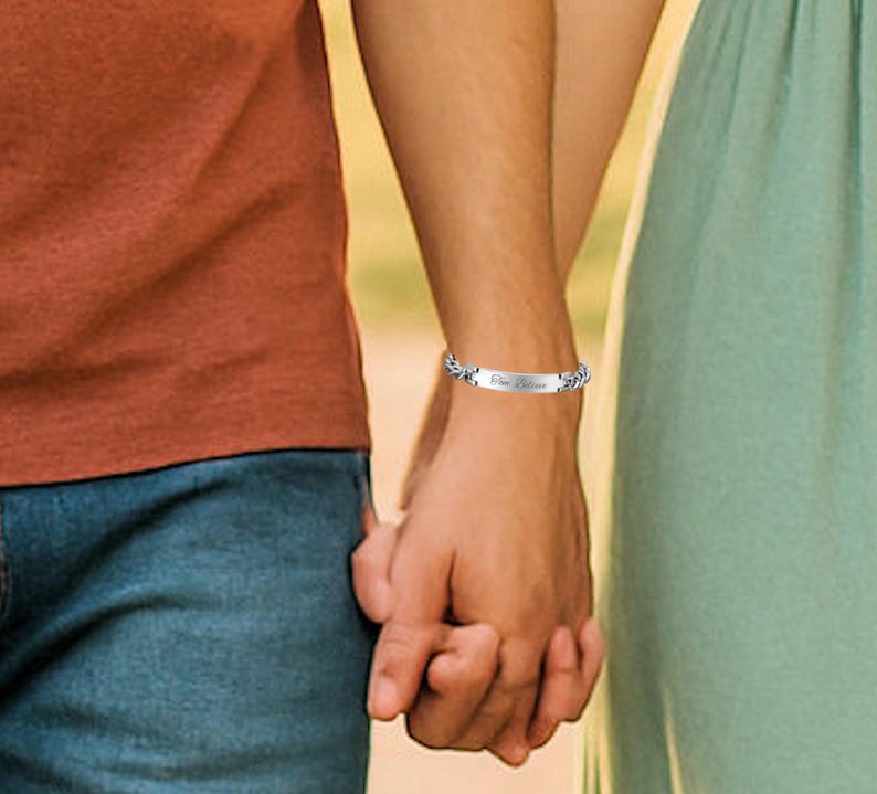 May include: A silver bracelet with the inscription "One Believe" is worn on a wrist. The bracelet has a decorative chain design. The background shows a person wearing an orange shirt and blue jeans holding hands with another person wearing a green dress.