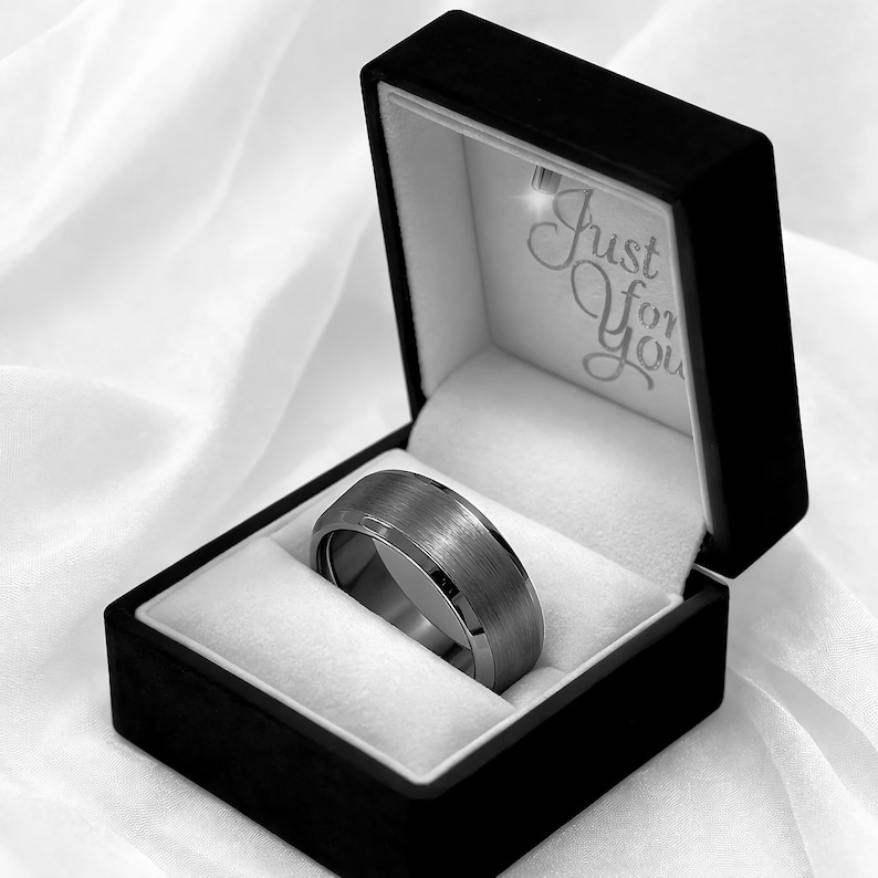 May include: A silver-colored ring with a brushed metal finish, presented in a black box with a white interior. The box is open, revealing the ring nestled on a white cushion. The text "Just For You" is visible inside the box.