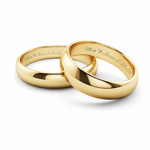 Puede incluir: Dos anillos de boda dorados, uno anidado dentro del otro. Cada anillo est&aacute; grabado con un nombre, un s&iacute;mbolo de coraz&oacute;n y una fecha. Los anillos tienen una superficie pulida y reflectante, sobre un fondo blanco.
