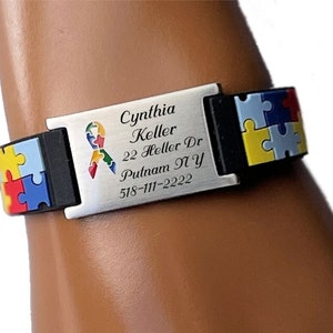 May include: A silver-colored medical ID bracelet with black accents and colorful puzzle piece designs. The bracelet has the name "Cynthia Keller", address, and phone number engraved on it. The bracelet is worn on a wrist.
