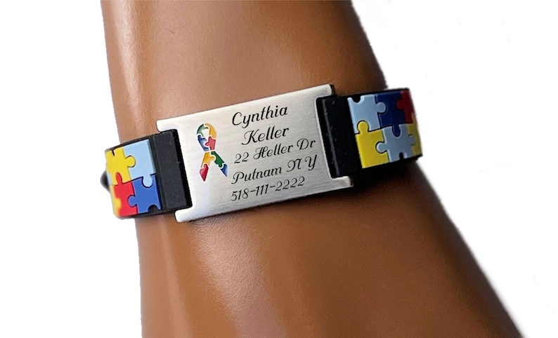 May include: A silver-colored medical ID bracelet with black accents and colorful puzzle piece designs. The bracelet has the name "Cynthia Keller", address, and phone number engraved on it. The bracelet is worn on a wrist.