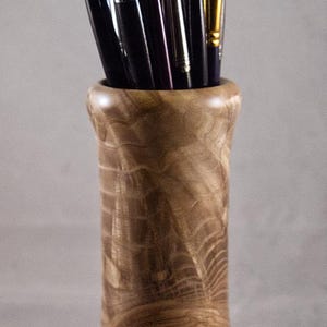 May include: A selection of paint brushes with black bristles and silver handles in a wooden vase. The vase is made of light brown wood with a natural grain pattern. The brushes vary in size and shape.