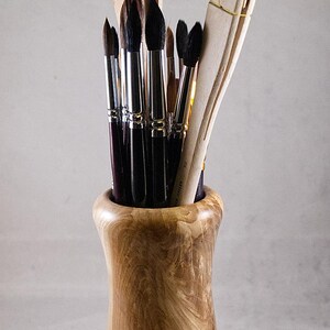 Peut inclure: Un pot en bois pour pinceaux d'artiste rempli de divers pinceaux. Le pot est en bois clair avec un motif de grain naturel. Les pinceaux ont des manches noirs et des poils de couleur claire. Certains pinceaux sont grands, d'autres petits.