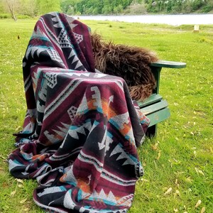 May include: A patterned blanket draped over a green wooden chair outdoors. The blanket features a geometric design in black, burgundy, gray, teal, and orange. The scene is set on a grassy area with a river and trees in the background.