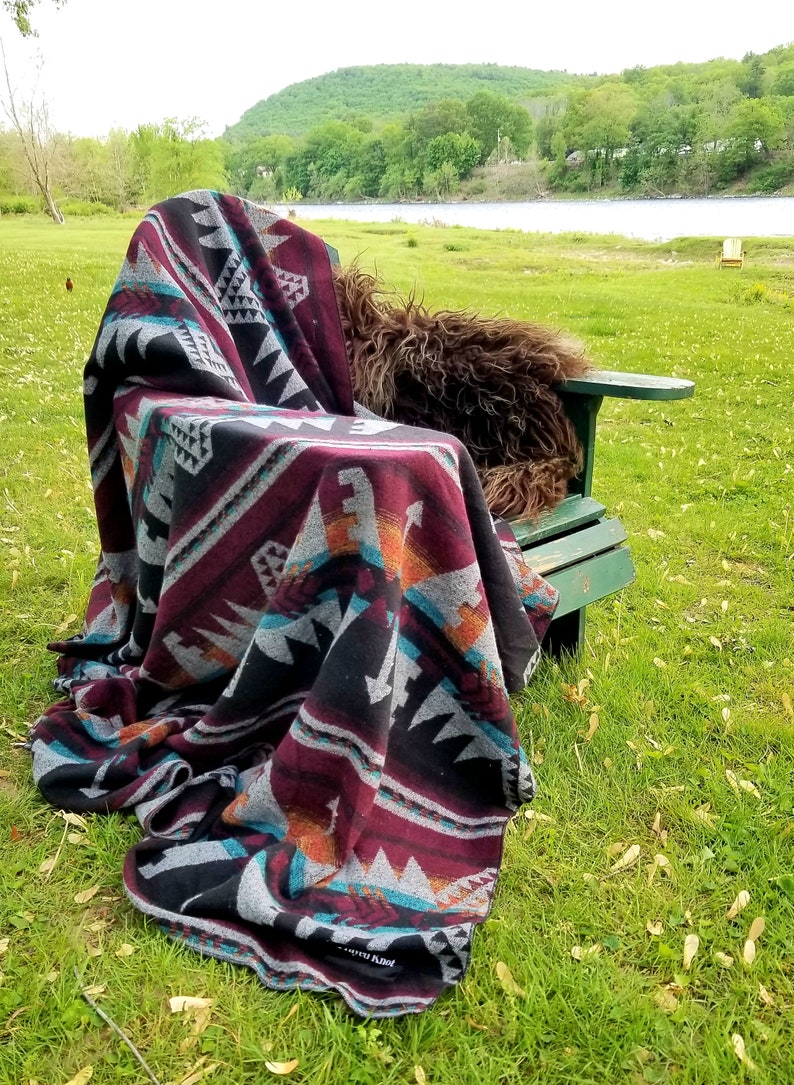 May include: A patterned blanket draped over a green wooden chair outdoors. The blanket features a geometric design in black, burgundy, gray, teal, and orange. The scene is set on a grassy area with a river and trees in the background.