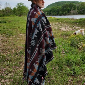May include: A person wearing a beige fedora and a patterned poncho with burgundy, black, gray, and turquoise geometric designs. The poncho is draped over the shoulders. The person is standing in a field of grass and flowers.