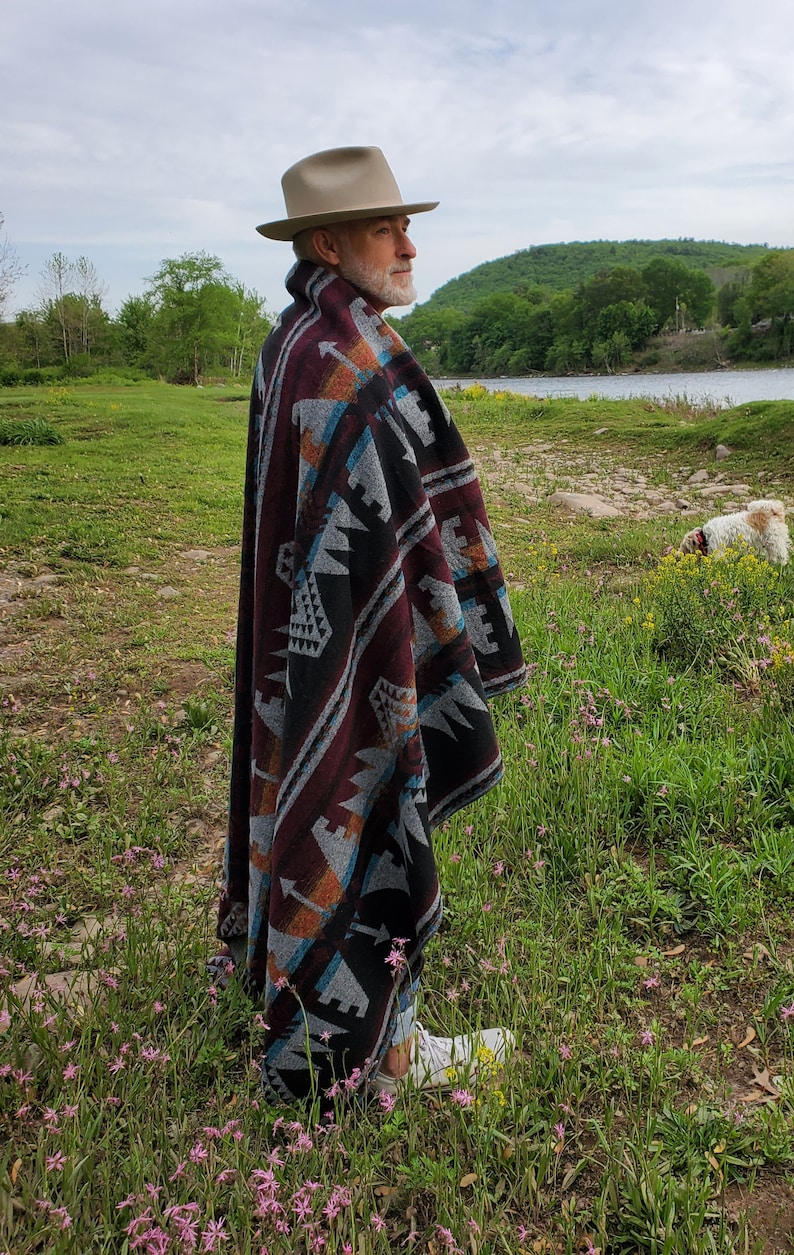 May include: A person wearing a beige fedora and a patterned poncho with burgundy, black, gray, and turquoise geometric designs. The poncho is draped over the shoulders. The person is standing in a field of grass and flowers.