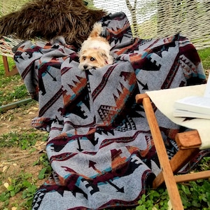 May include: A gray blanket with a geometric pattern in shades of black, orange, and burgundy is draped over a hammock. A small dog is nestled in the blanket. A wooden chair and a book are in the background. The blanket is made of a soft material.