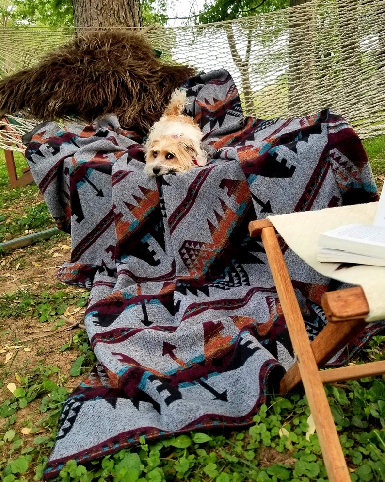 May include: A gray blanket with a geometric pattern in shades of black, orange, and burgundy is draped over a hammock. A small dog is nestled in the blanket. A wooden chair and a book are in the background. The blanket is made of a soft material.