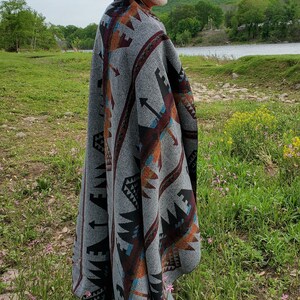 May include: A gray blanket with a geometric pattern in shades of brown, black, and teal. The blanket is draped over the shoulders. The person is wearing a tan hat. The scene is outdoors with a river and trees in the background.