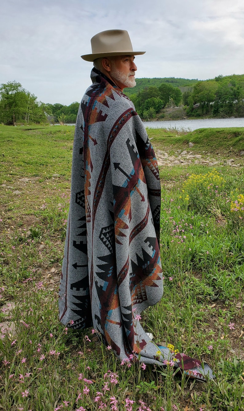 May include: A gray blanket with a geometric pattern in shades of brown, black, and teal. The blanket is draped over the shoulders. The person is wearing a tan hat. The scene is outdoors with a river and trees in the background.
