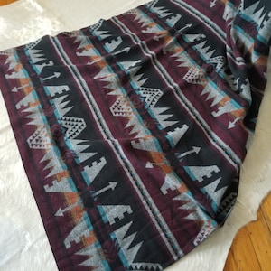 May include: A burgundy, black, and gray patterned blanket with geometric and arrow designs. The blanket has stripes of varying widths and colors, including teal and orange. The blanket is folded and draped over a white surface.