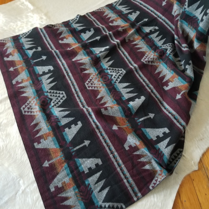 May include: A burgundy, black, and gray patterned blanket with geometric and arrow designs. The blanket has stripes of varying widths and colors, including teal and orange. The blanket is folded and draped over a white surface.