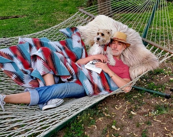May include: A person relaxing in a white rope hammock with a dog and a blanket. The blanket has a red, teal, and brown geometric pattern. The person is wearing a red shirt, blue jeans, and a straw hat. A white fluffy rug is behind them.