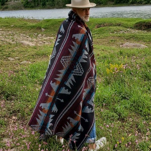 May include: A person wearing a beige fedora hat and a patterned blanket with black, burgundy, and teal geometric designs. The person is walking in a field of pink flowers near a river.