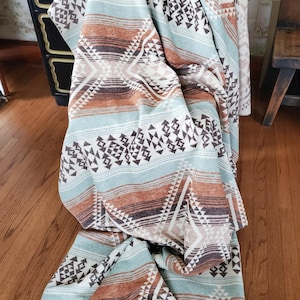 May include: A folded blanket with a Southwestern geometric pattern in shades of teal, brown, and cream. The blanket is draped over a dark dresser and wooden chair, showcasing its design and texture. The blanket's pattern includes triangles and stripes.
