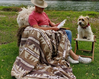 May include: A brown and beige patterned blanket with geometric designs. The blanket is draped over a chair. A person is sitting in the chair, wearing a red shirt, blue jeans, and a beige hat. A small dog sits on a folding chair nearby.