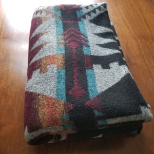 May include: Folded blanket with a Southwestern design. The blanket features a pattern in shades of gray, black, teal, burgundy, and orange. The design includes geometric shapes and arrow-like motifs, creating a textured appearance.