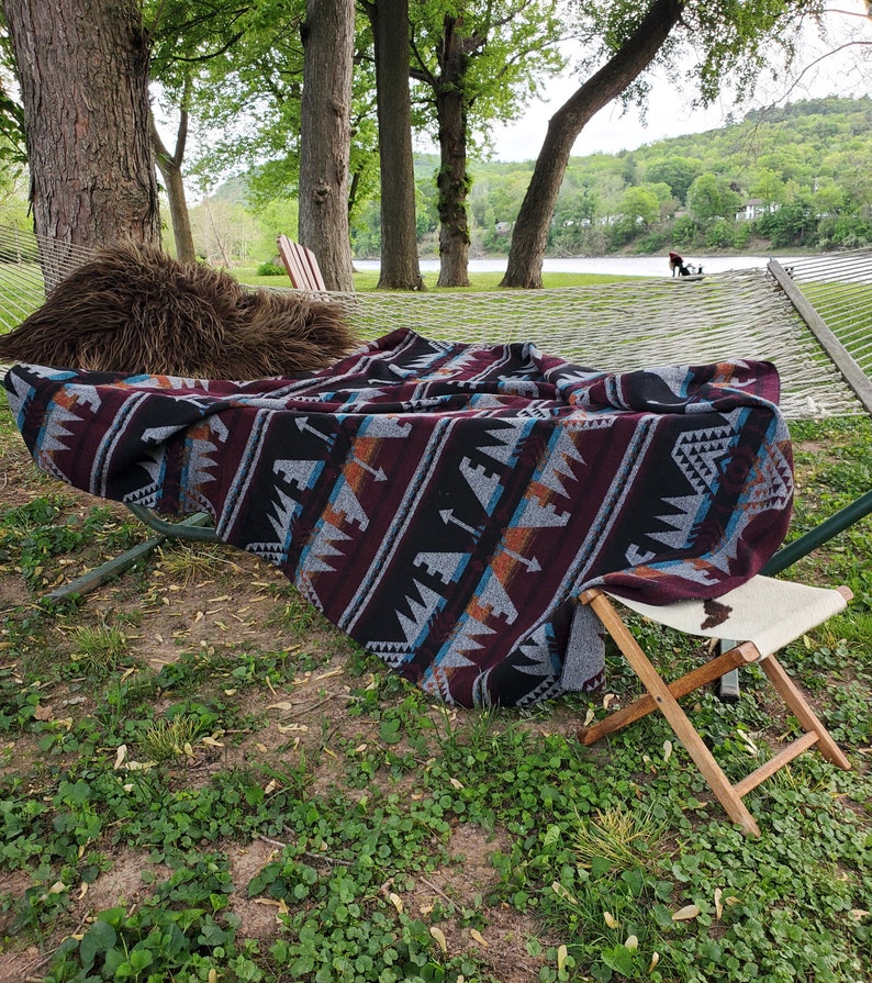 May include: A patterned blanket with a burgundy, black, and gray design is draped over a hammock. A small folding wooden stool with a white seat is next to the hammock. A brown fur pillow rests on the hammock.