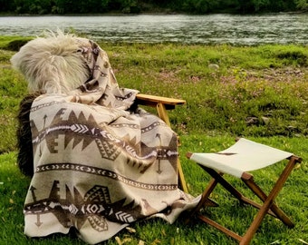 May include: A wooden folding chair with a beige and brown patterned blanket draped over it, and a small folding stool, set on a grassy area near a river. The blanket features geometric designs. A fluffy, light-colored cushion rests on the chair.
