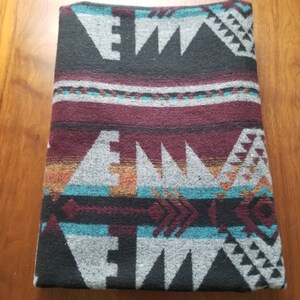 May include: Folded blanket with a Southwestern geometric pattern. The blanket features a design with gray, black, burgundy, teal, and orange colors. The pattern includes triangles and arrow shapes.