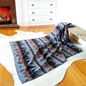 May include: A gray woven blanket with a Southwestern pattern in shades of burgundy, orange, and teal. The blanket is draped over a wooden bench and a white cowhide rug on a hardwood floor.
