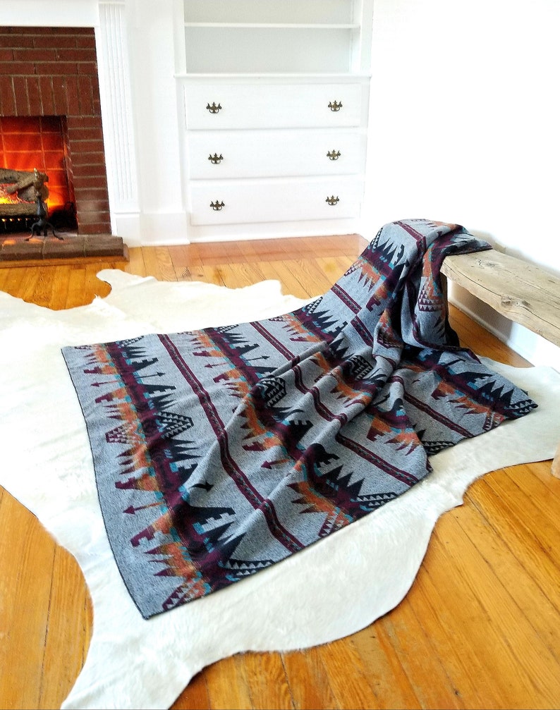 May include: A gray woven blanket with a Southwestern pattern in shades of burgundy, orange, and teal. The blanket is draped over a wooden bench and a white cowhide rug on a hardwood floor.