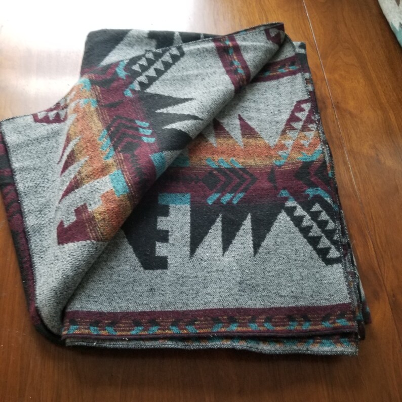 May include: A folded gray blanket with a Southwestern geometric pattern. The design features black, burgundy, teal, and orange accents. The blanket is folded over, revealing the patterned design.
