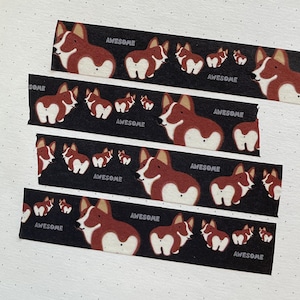 Può includere: Cinque rotoli di nastro washi nero e bianco con un motivo ripetuto di corgi marroni e bianchi. Il testo "AWESOME" è stampato su ogni rotolo.