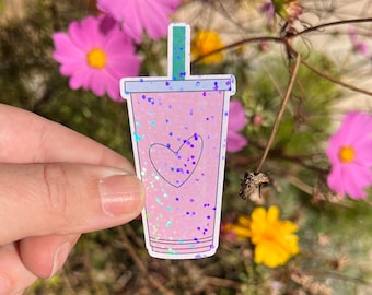 May include: Pink glitter sticker of a pink cup with a straw and a heart on the side. The sticker is on a hand in front of a blurry background of pink and yellow flowers.