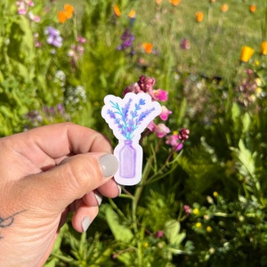 May include: A purple watercolor sticker of a bouquet of lavender flowers in a clear glass bottle. The sticker is held in a hand against a blurred background of a field of flowers.
