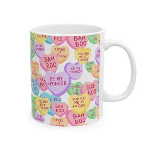 May include: White ceramic mug featuring a repeating pattern of colorful heart-shaped candies. Each candy has a unique message, including phrases like "BAH BOO" and "BE MY SPONSOR". The mug has a curved handle.