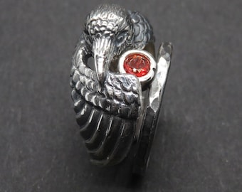 May include: A silver ring with a detailed bird design and a red gemstone. The bird's wings are spread out and the gemstone is set in the center of the ring.