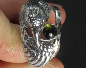 May include: A silver ring with a detailed bird design and a green gemstone. The bird's head is turned to the side and the gemstone is set in the center of the ring.